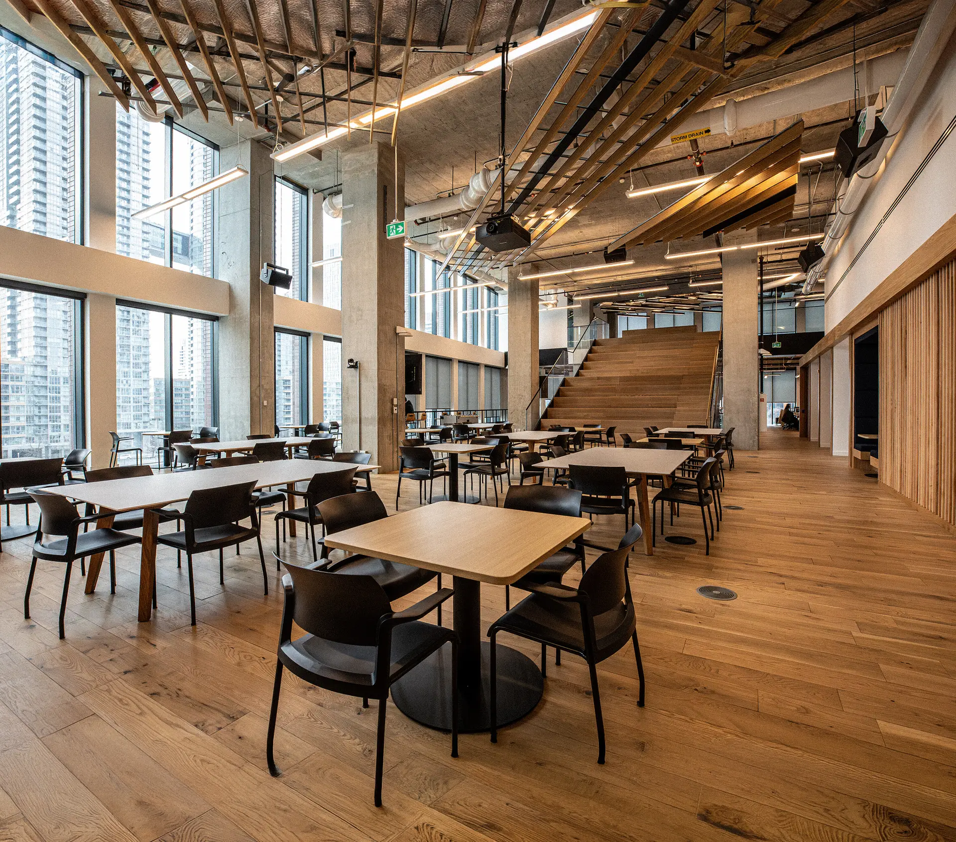 Office dining area with communal tables beside floor-to-ceiling windows overlooking the city