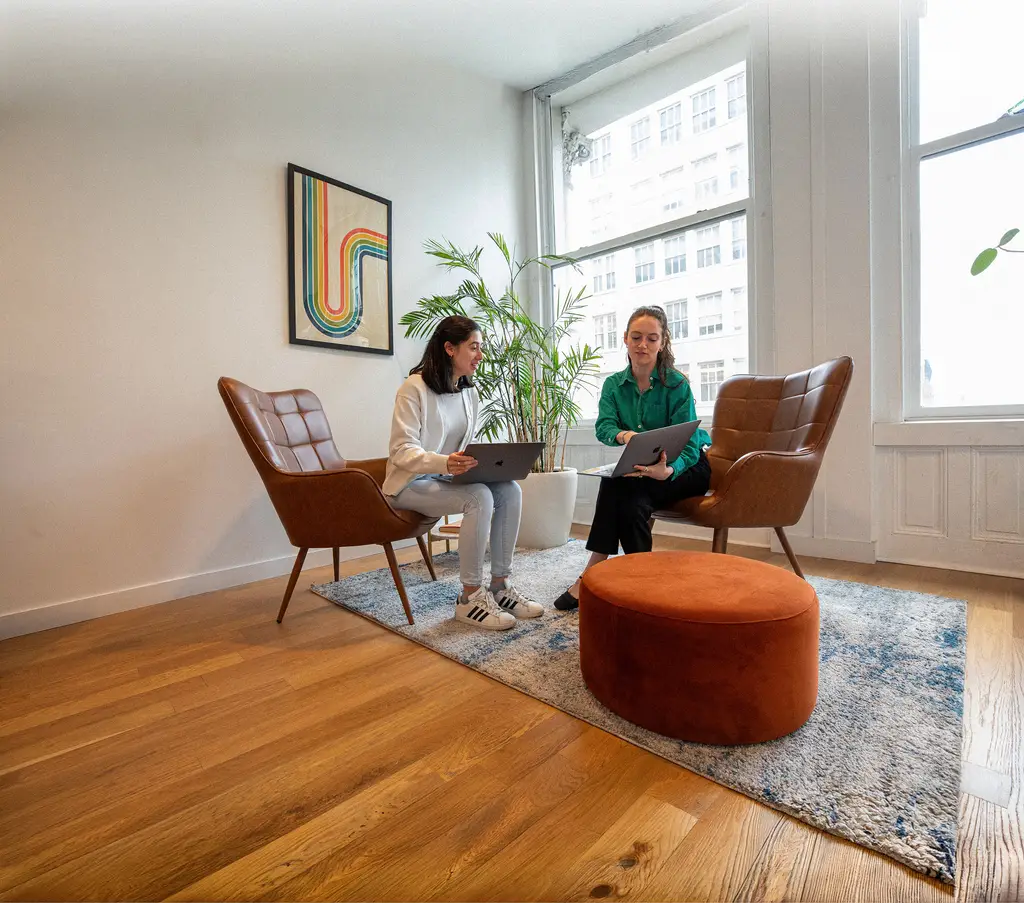 Two Konrad team members reviewing work on laptops in leather lounge chairs beside a sunlit window
