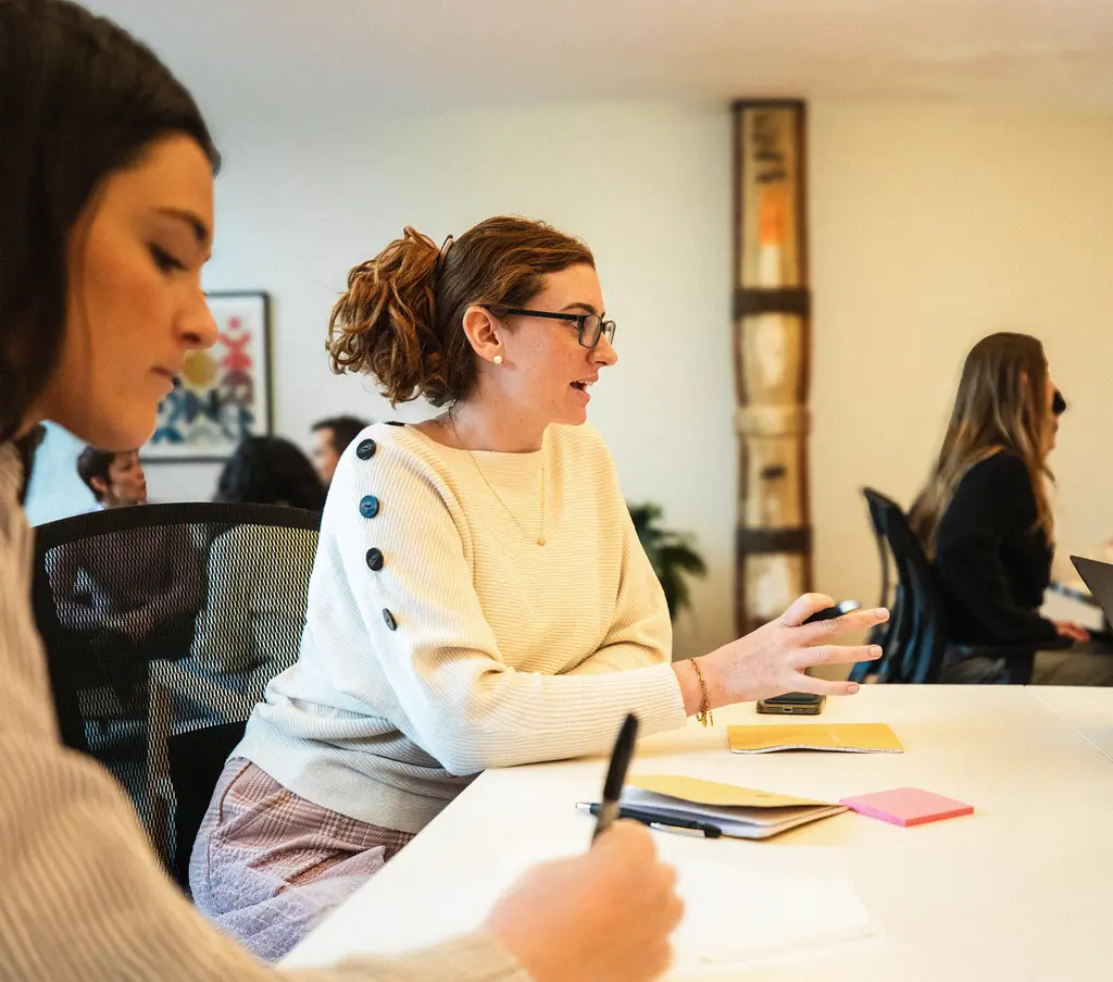 Two team members in conversation at a meeting table, one gesturing as she speaks