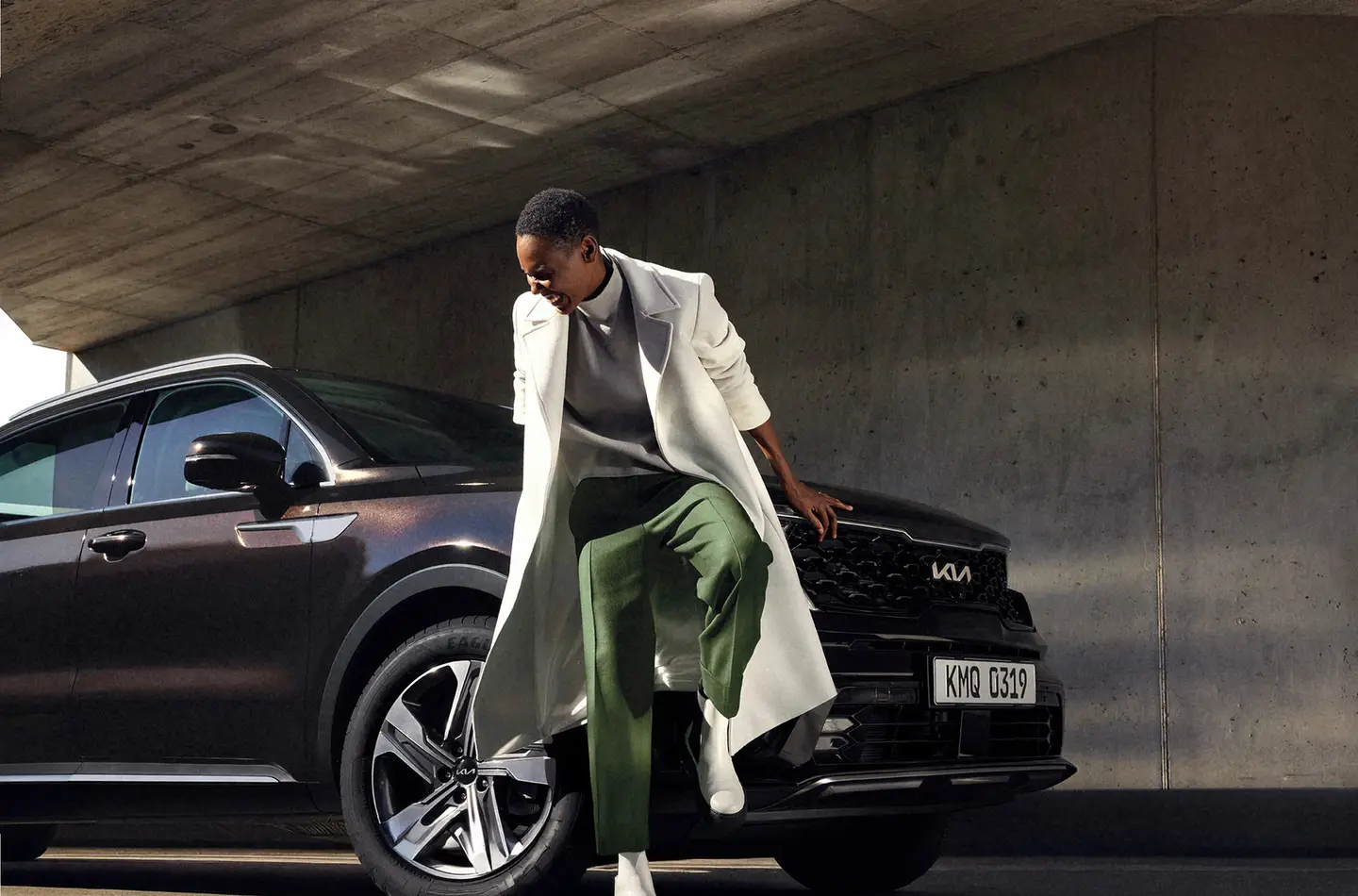 Person in a long coat leaning against the hood of a dark Kia in a concrete garage