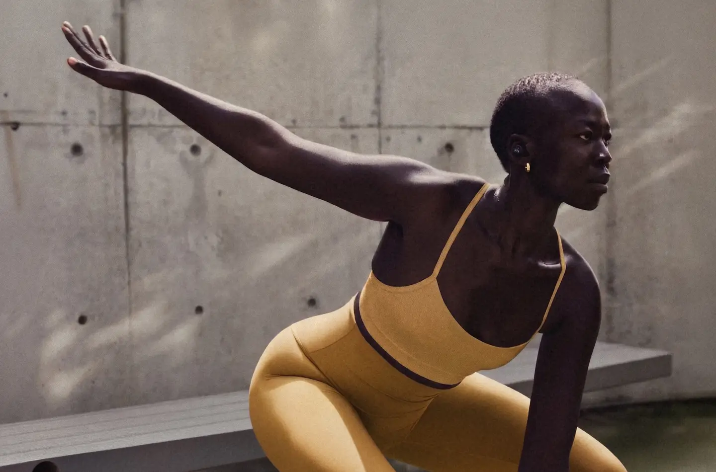 Close-up of an athlete’s shoulder in a yellow sports bra, wearing a wireless earbud, against a concrete wall