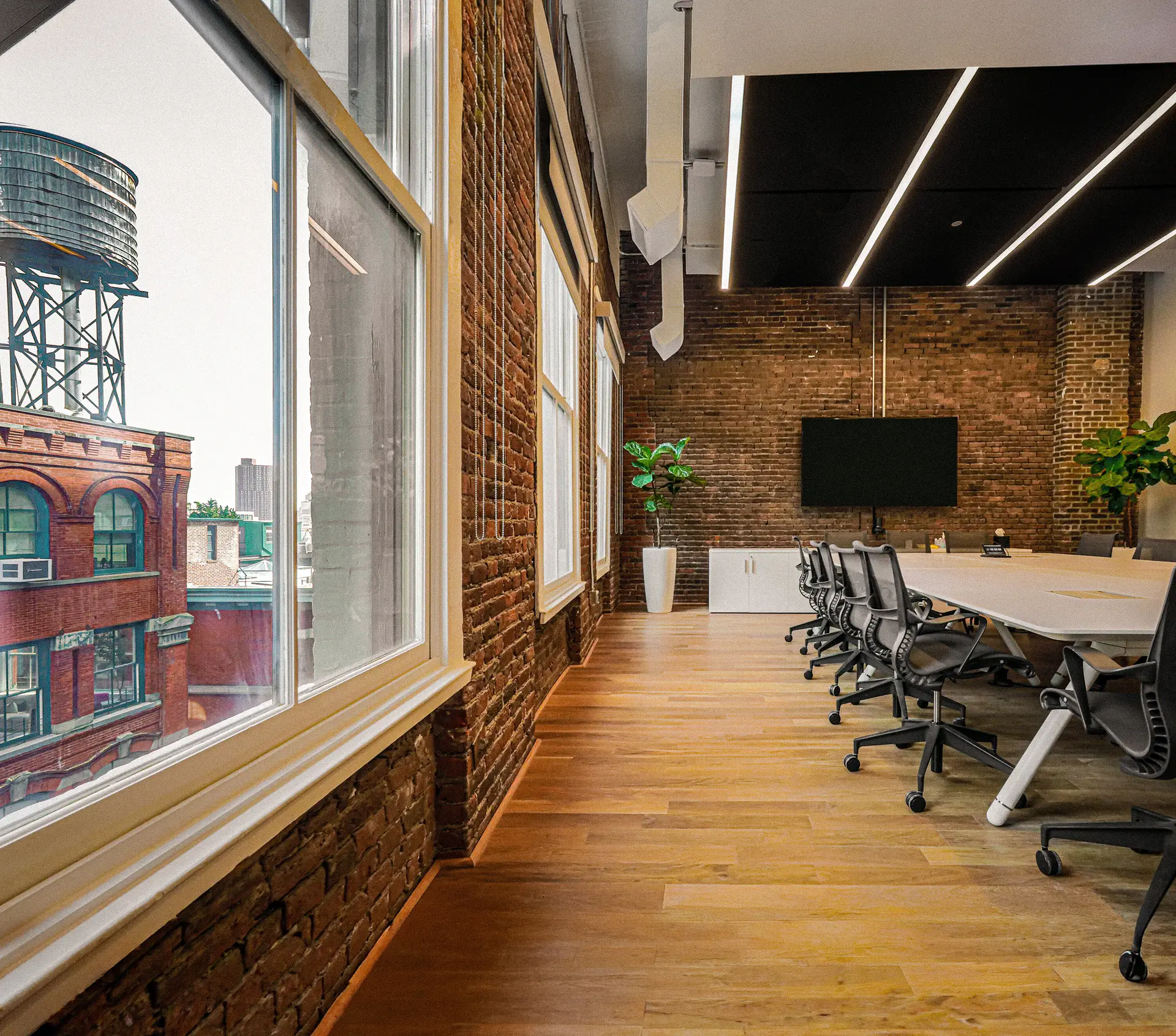 Sunlit office boardroom with an exposed brick wall, a long meeting table, and tall windows overlooking historic brick buildings and a rooftop water tower